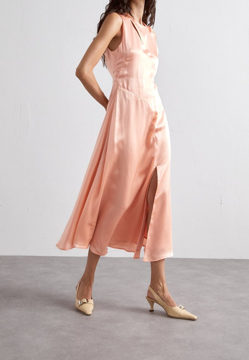 Satin midi-klänning i ljus persika med ett sidoslit och ärmlös design. Kombinerad med beige slingback-klackar med metallisk accent.