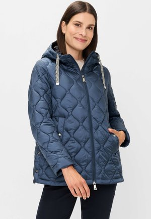 Frau trägt eine gesteppte marineblaue Kapuzenjacke mit gemusterten Kordeln, Hände in den Taschen, leicht lächelnd, vor einem schlichten Hintergrund.