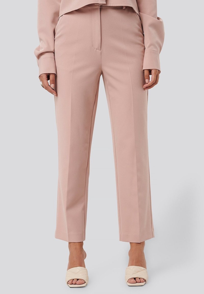 Femme portant un pantalon taille haute à jambes droites rose poudré avec des plis avant et des talons ouverts crème matelassés sur un fond clair.