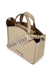 Beige ādas rokas soma ar strukturētu dizainu, izliektiem malām, melniem akcentiem un noņemamu siksnu, kurā ar lieliem burtiem ir rakstīts 'KARL LAGERFELD'.