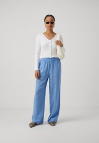 Un cardigan blanc à côtes avec des manches longues, un pantalon décontracté bleu clair, et un sac à main crème ; les accessoires incluent des lunettes de soleil noires et des chaussures beiges.