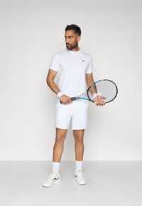 Λευκό αθλητικό μπλουζάκι και σορτσάκι, με πράσινο λογότυπο. Φοριούνται με λευκά κάλτσες και αθλητικά παπούτσια. Κρατώντας μία ρακέτα τένις. Καθαρός σχεδιασμός.