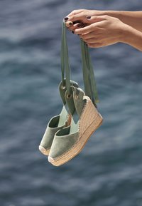 Green suede espadrilles with a woven jute base, featuring two fabric straps and grommets for hanging. Smooth texture and rounded toe design.
