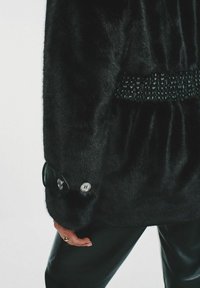 Czarna kurtka z sztucznego futra z marszczoną talią i teksturowanym czarnym detalem. Posiada błyszczący czarny guzik z srebrnym akcentem.