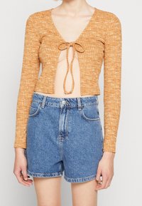 Orange långärmad cropped topp med knytdekoration framtill, kombinerad med högmidjade blå denimshorts. Texturerad tyg, avslappnad passform.