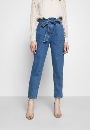Femme portant un jean bleu taille haute avec une ceinture en tissu nouée, associé à des talons beiges à bout ouvert et un chemisier crème à manches longues.