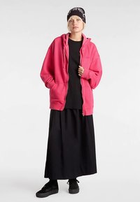 Roze hoodie met rits van fleece materiaal met een buidelzak, gedragen over een zwart T-shirt en een lange zwarte rok. Zwarte sneakers maken de look compleet.