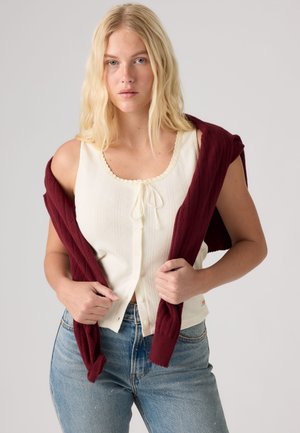 Femme blonde portant un haut blanc sans manches boutonné avec une cravate et un pull bordeaux drapé sur ses épaules.