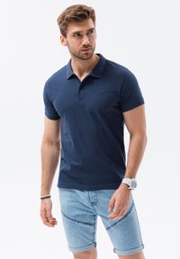 Marinblå polo skjorta med krage och korta ärmar, med en diskret broderad logotyp. Matchad med ljusa denimshorts och en klocka.