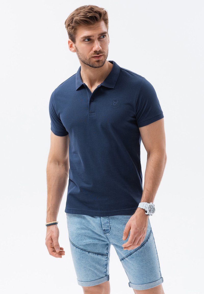 Marinblå polo skjorta med krage och korta ärmar, med en diskret broderad logotyp. Matchad med ljusa denimshorts och en klocka.