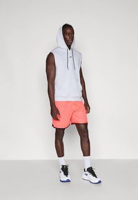 Moški stoji oblečen v brezrokavnik sivo barve, svetle koralne športne kratke hlače, bele nogavice in bele superge z modrimi poudarki.