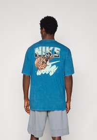 Türkiissinine Nike'i T-särk, mille seljal on julged mustad ja korallpunased graafikud korvpallist ja sõnast "BASKETBALL". Lühikesed varrukad, puuvillane materjal.