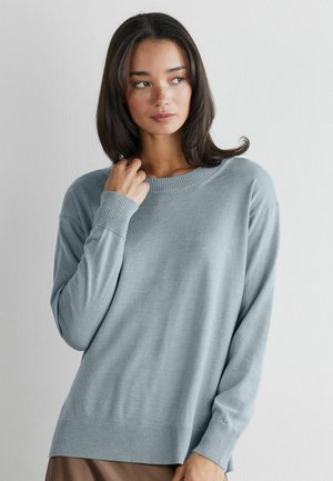 Femme aux cheveux bruns mi-longs portant un pull en tricot bleu clair, regardant légèrement sur le côté, la main touchant l'encolure sur un fond uni.