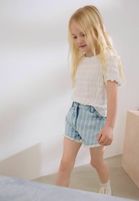 Witte getextureerde top met korte mouwen en lichtblauwe gestreepte denim shorts met rafelige randen. De achtergrond heeft neutrale tinten en een minimalistisch ontwerp.