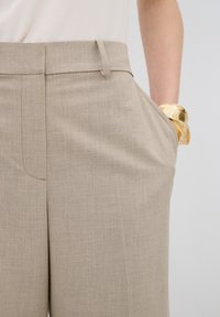 Beige kiilatud püksid tekstureeritud kangaga, slim fit ja küljetaskutega. Kuldsed käevõrud on nähtavad randmel.