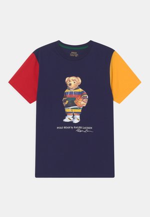 T-shirt bleu marine avec des manches courtes rouges, jaunes et vertes. Arbore un graphique d'un ours en tenue de sport tenant un ballon de basket. Matière en coton.
