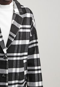 Blazer a quadri bianco e nero con un design strutturato, caratterizzato da grandi bottoni e un colletto a revers. Realizzato in tessuto misto lana.