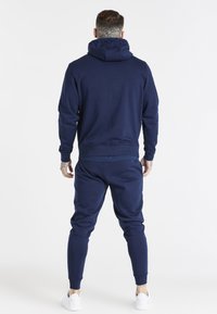 Navy blue, cotton hoodie with a drawstring hood, paired with matching tapered sweatpants. White sneakers visible. Smooth texture throughout.