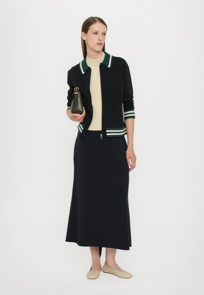 Cardigan negru cu guler în dungi verzi și albe, top bej, fustă neagră și balerini de culoare deschisă, ținând o geantă mică neagră.
