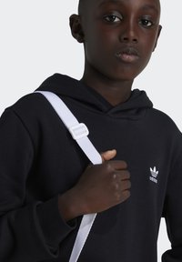 Svart huvtröja med en vit Adidas-logotyp på bröstet, med ett vitt band och spänne över axeln. Slät tygtextur.