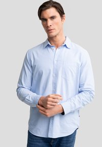 Mann, der den Ärmel einer hellblauen Hemdjacke justiert, steht vor einem einfachen grauen Hintergrund.