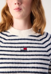 Stripet genser laget av mykt, teksturert garn; har horisontale marineblå striper på en kremfarget bakgrunn og et lite "TOMMY JEANS" logo-patch.