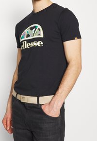 Černé bavlněné tričko s multikolorovým maskáčovým logem a zlatými akcenty. Nosíno s tmavými džíny a béžovým opaskem.