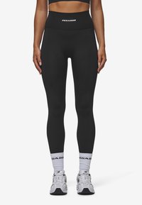 Zwarte high-waisted leggings met een gladde, rekbare textuur en het "PEGADOR" logo op de tailleband, gecombineerd met witte sokken en sneakers.