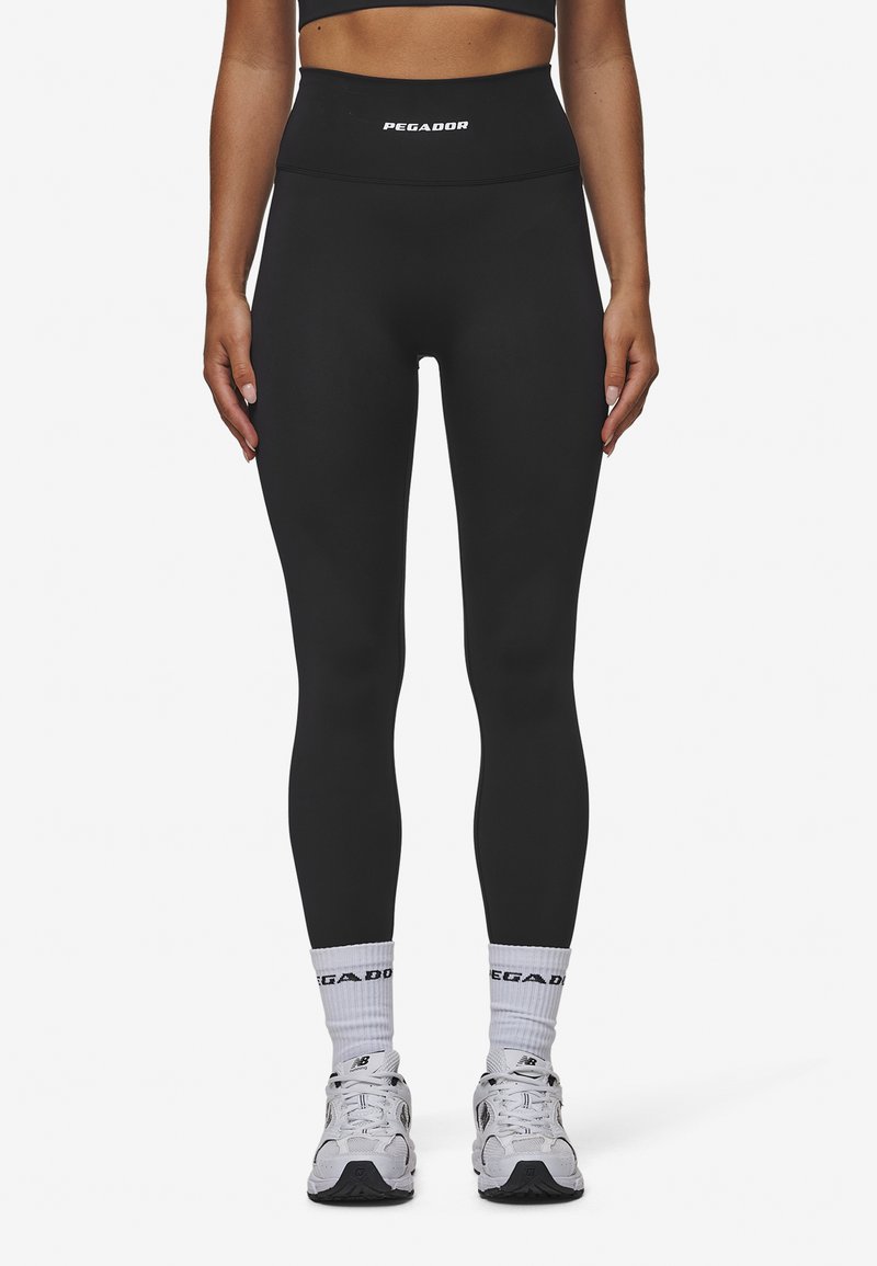 Zwarte high-waisted leggings met een gladde, rekbare textuur en het "PEGADOR" logo op de tailleband, gecombineerd met witte sokken en sneakers.
