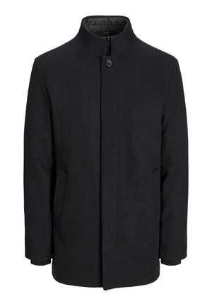 Man draagt een zwarte jas met knopen over een bruine trui met rits en een wit overhemd met kraag, grijze broek en handen in de jaszakken.