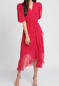 Frau in einem roten Midi-Wickelkleid mit weißen Punkten, puffförmigen Ärmeln, rüschigem Saum und beigen High-Heels.