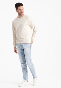 Felpa beige con colletto tondo, abbinata a jeans slim-fit chiari e sfrangiati e sneaker bianche. Tessuto morbido con polsini a costine.
