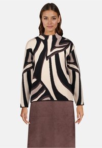 Vrouw draagt een trui met lange mouwen met zwart, beige en bruin abstract patroon en een hoge hals, gecombineerd met een bruine rok.