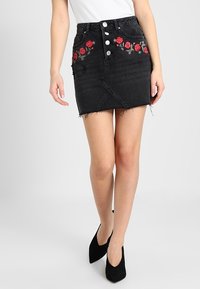 Svart denimkjol med broderade röda rosor, knappstängning, fransad fåll och asymmetrisk design. Bärs med svarta klackar.