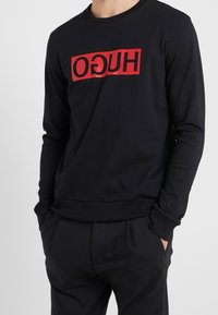 Hombre vestido con una camisa de manga larga negra, con un logo rectangular rojo y el texto "HUGO" en reverso, con las manos en los bolsillos, frente a un fondo gris liso.