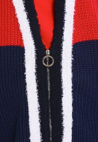 Stickad kofta med röda, marinblå och vita färgblock, med dragkedja och texturerade stickningsmönster genom hela plagget.