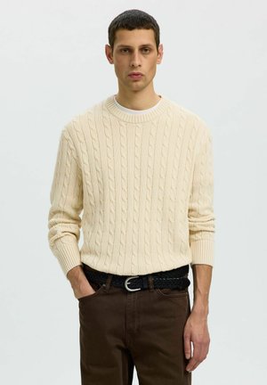 Jeune homme aux cheveux courts et foncés portant un pull torsadé crème, un pantalon marron et une ceinture tressée noire, debout avec une main dans la poche.