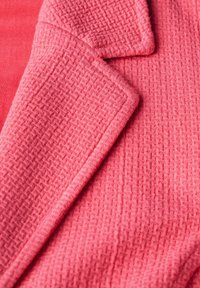 Materiał tekstylny w kolorze koralowym o fakturze z plecionym wzorem, z wyprofilowanym kołnierzem i wyraźnymi krawędziami, cechujący się miękkim, ciepłym wyglądem.
