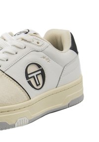 Scarpa sportiva bianca realizzata in pelle e suede, con un logo rotondo, accenti neri e una suola testurizzata con una intersuola beige.