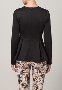 Top peplum noir à manches longues, en tissu texturé, avec une fermeture éclair au dos. Porté avec un pantalon à motifs multicolores présentant des éclaboussures de couleur.