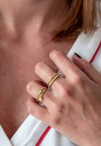 Arany színű gyűrűk csavart mintával, az egyik csillag díszítéssel, a másik zöld szív részlettel, egy kézen viselve.