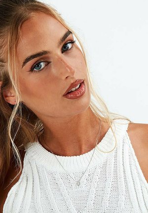White knitted sleeveless top with a ribbed texture. A fine silver necklace is visible, complementing the skin tone and hair.