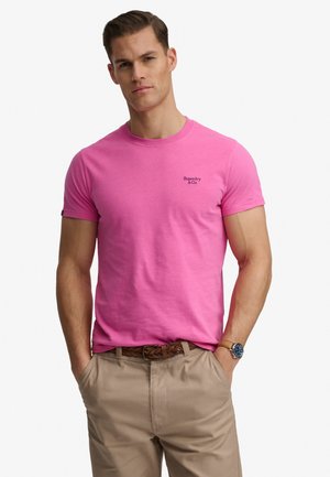 Hombre con camiseta rosa de manga corta y cuello redondo con el logo "Superdry & Co", pantalones beige, cinturón tejido y reloj de pulsera, manos en los bolsillos.