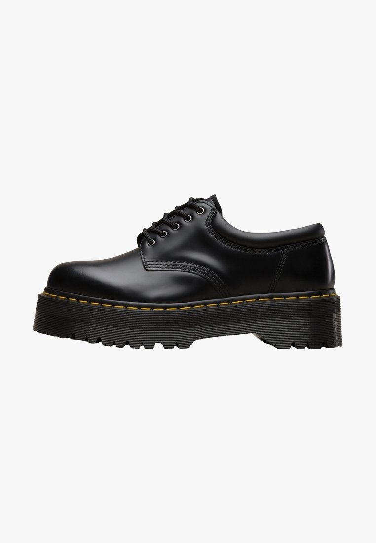Chaussure en cuir noir à lacets avec une semelle épaisse à rainures et une couture jaune le long du bord, vue de côté sur fond blanc.