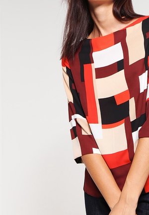 Kvinde iført en top med geometrisk mønster med røde, beige, sorte og hvide felter, med ærmer til albuen og bådudskæring.