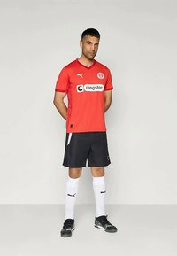 Άνδρας που στέκεται με τα χέρια πίσω από την πλάτη, φορόντας κόκκινη φανέλα ποδοσφαίρου Puma, μαύρες βερμούδες, λευκές κάλτσες και μαύρες ποδοσφαιρικές μπότες σε ουδέτερο φόντο.