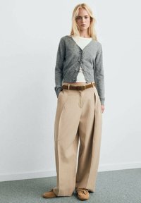 Cardigan gris avec des boutons ronds, associé à un pantalon beige àjambe large et une ceinture marron. Stylé avec un haut blanc et des chaussures marron.