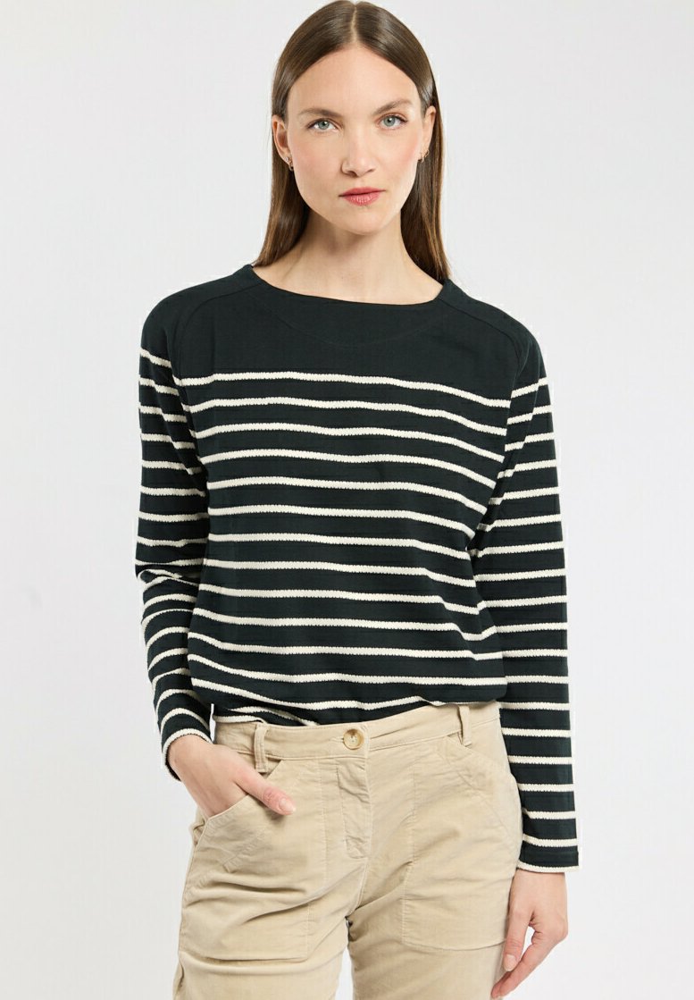 Frau mit langen, glatten braunen Haaren, die ein schwarz-weiß gestreiftes Langarmshirt und beige Hosen trägt, steht mit einer Hand in der Tasche.
