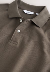 Polo de punto texturizado marrón con cuello extendido y dos botones blancos, etiquetado como corte regular, tamaño mediano.