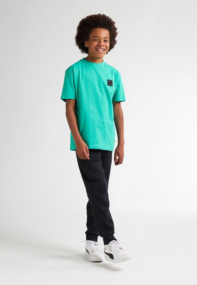 Turkis kortærmet t-shirt med en rund halsudskæring og et sort cirkulært logo patch. Sættes sammen med sorte sweatpants og hvide sneakers.